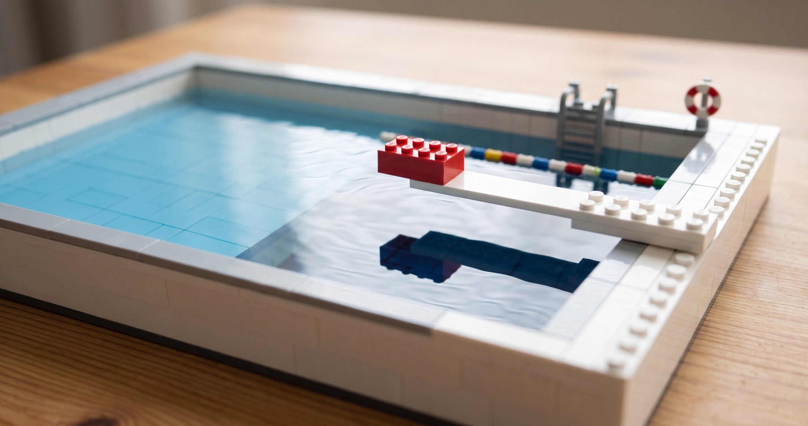 A small swimming pool built from toy bricks, with a red brick at the tip of a diving board over the deep end, and a ladder, a lifebuoy and a lane rope around it
